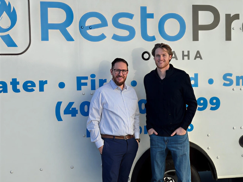 Two men standing and smiling in front of a white truck with blue and black text.