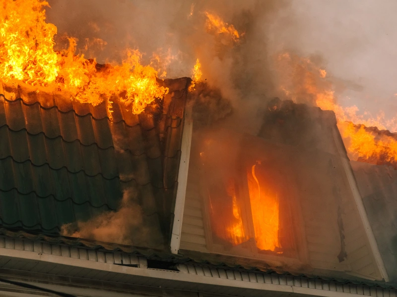 Flames and smoke engulf the roof and window of a burning house.