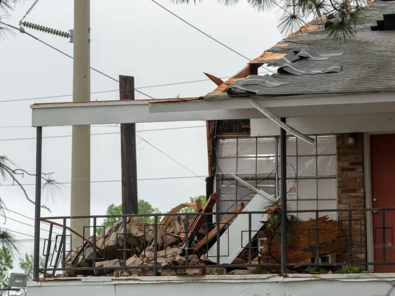 Damaged house porch with broken roof shingles and debris on the floor.