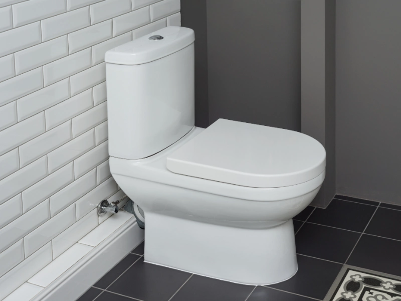 White modern toilet with closed lid in a bathroom with white tiled wall and dark floor tiles.