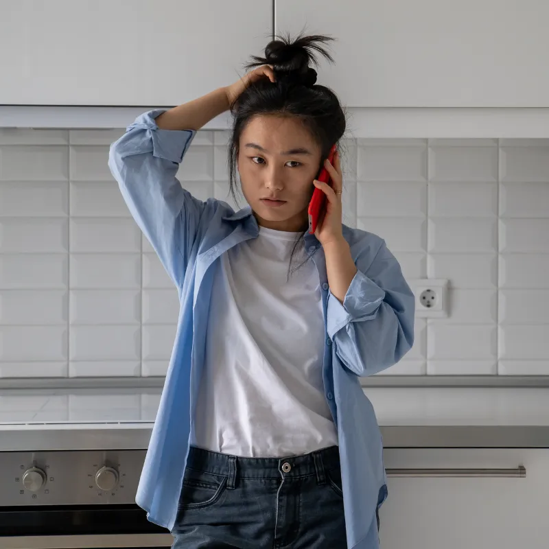Young woman in a blue shirt holding a red phone to her ear, looking concerned in a kitchen.